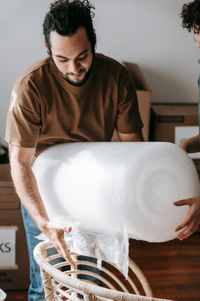 services-03 Focused ethnic man with roll of bubble wrap preparing to cover wicker armchair during apartment renovation in living room with anonymous partner
